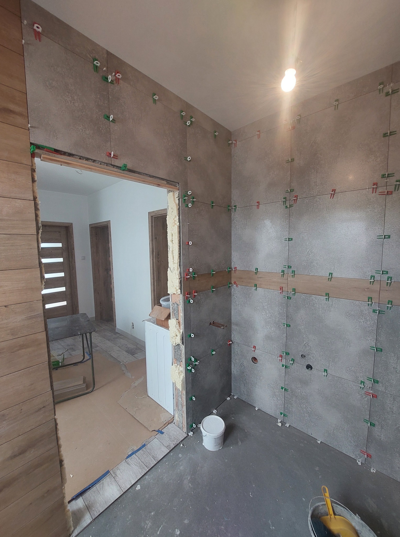 Układanie płytek w łazience, widoczne krzyżyki dystansowe. Częściowo wykończona ściana z szarych płytek i drewnopodobna okładzina przy drzwiach. Na podłodze wiadro i resztki zaprawy.