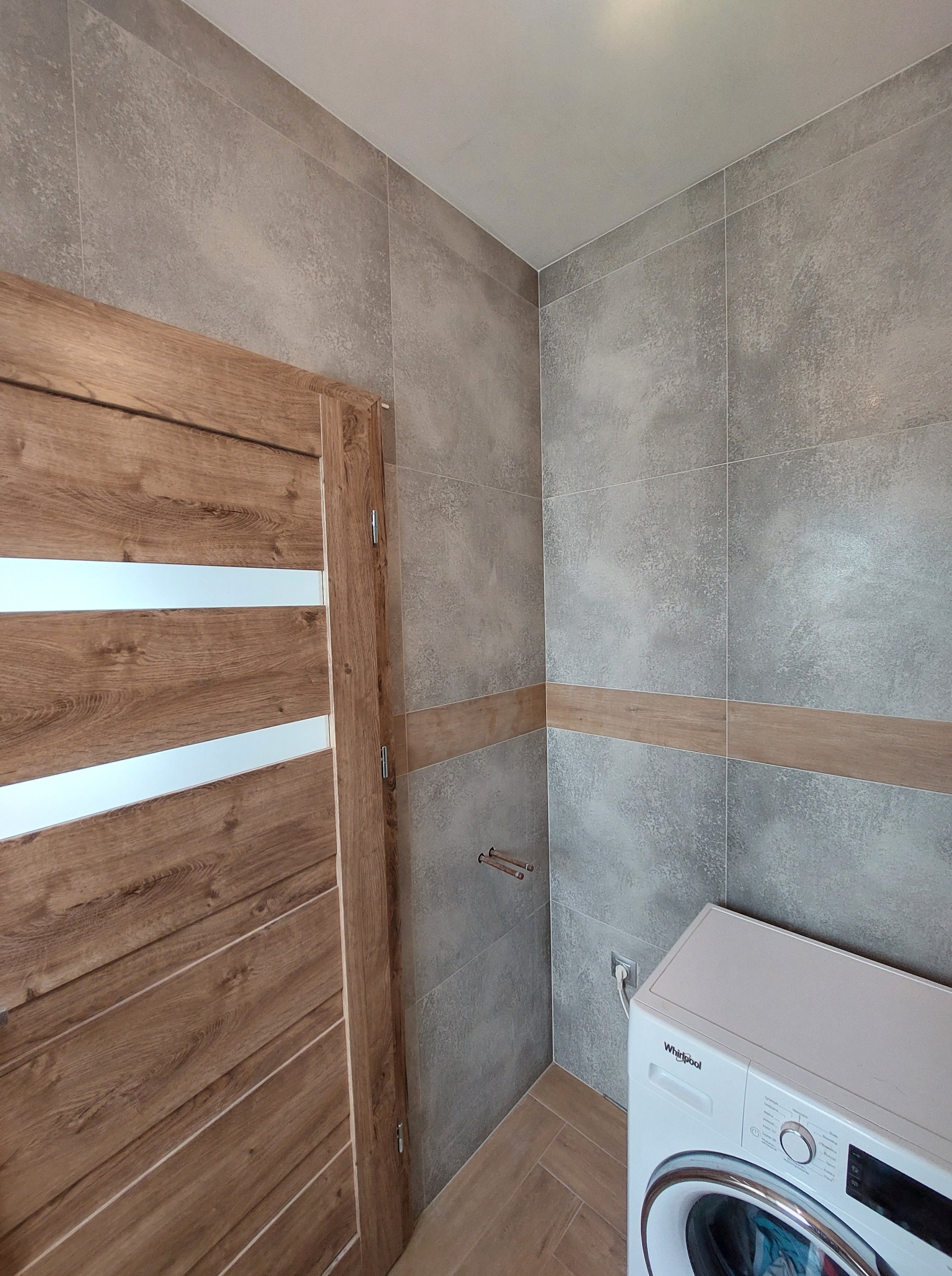 Wykończona łazienka z szarymi płytkami i drewnianymi akcentami. Widoczna pralka Whirlpool i nowoczesne drzwi z poziomymi przeszkleniami.