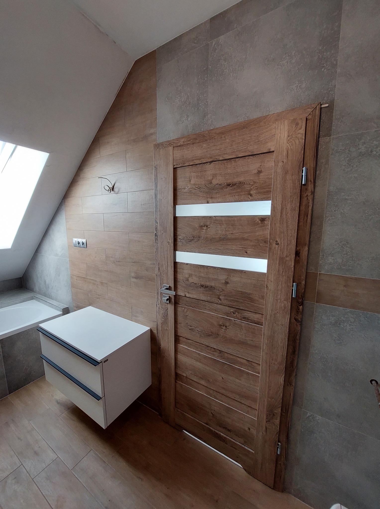 Wykończona łazienka na poddaszu: drewnopodobne płytki, biała szafka pod umywalkę z szufladami i drewniane drzwi z przeszkleniami, skośny sufit z oknem.