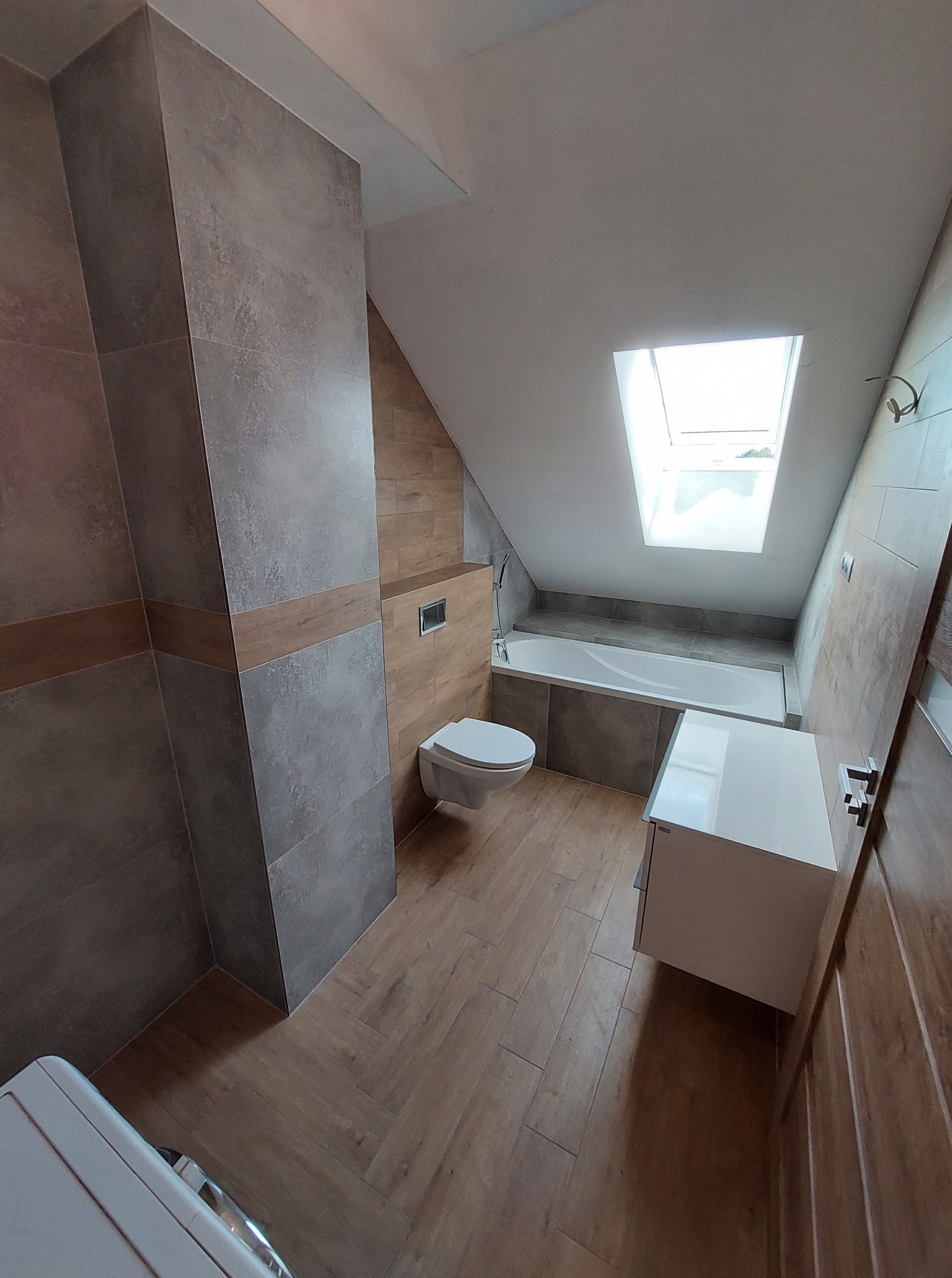 Jasna łazienka na poddaszu z oknem dachowym. Szare płytki ścienne z drewnianymi akcentami, drewniana podłoga w jodełkę, biała szafka pod umywalkę i toaleta.