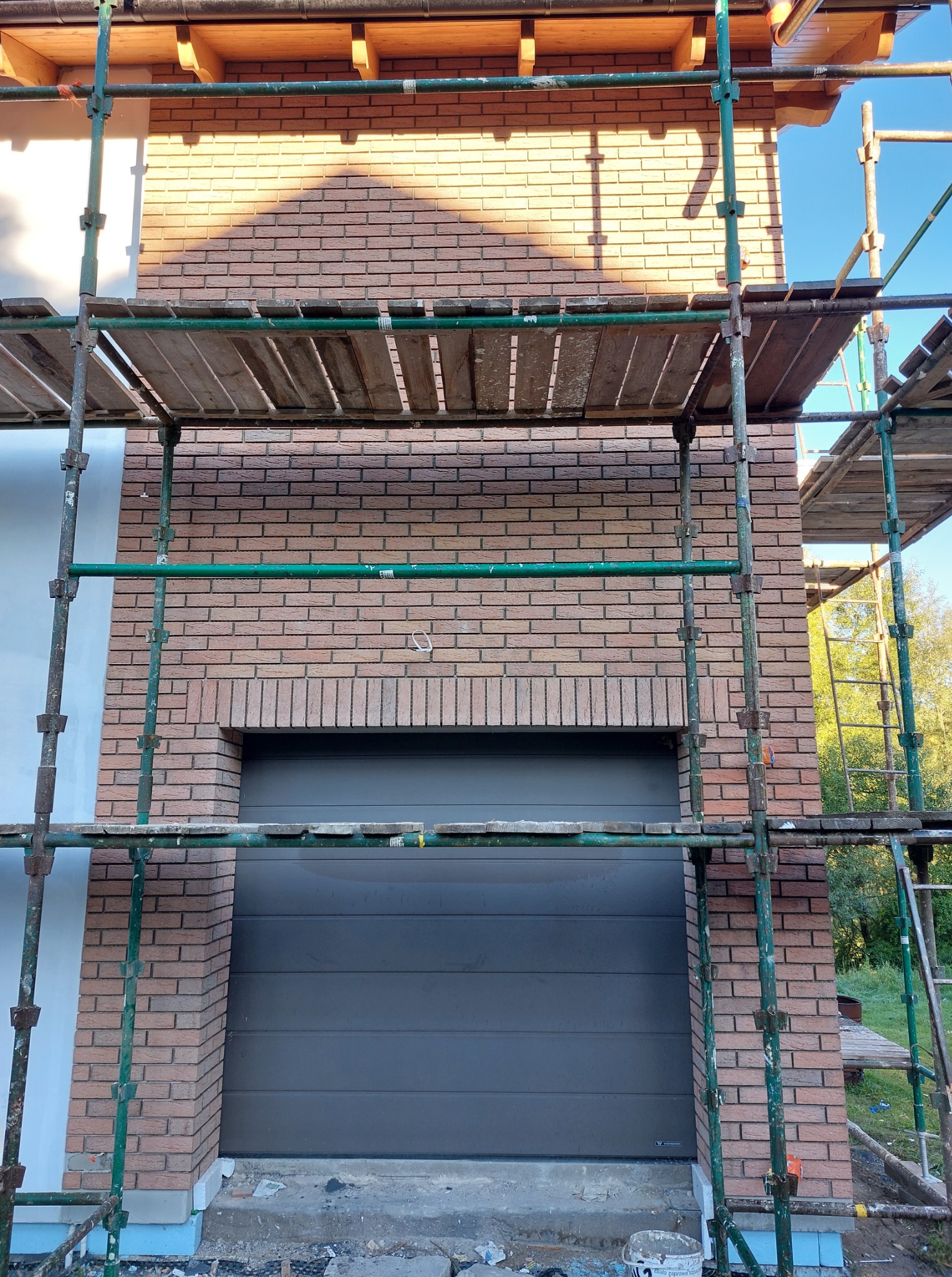 Murowanie elewacji z cegły klinkierowej w Sierakowicach. Widoczne rusztowanie i brama garażowa. Cieniowanie na elewacji dodaje głębi.