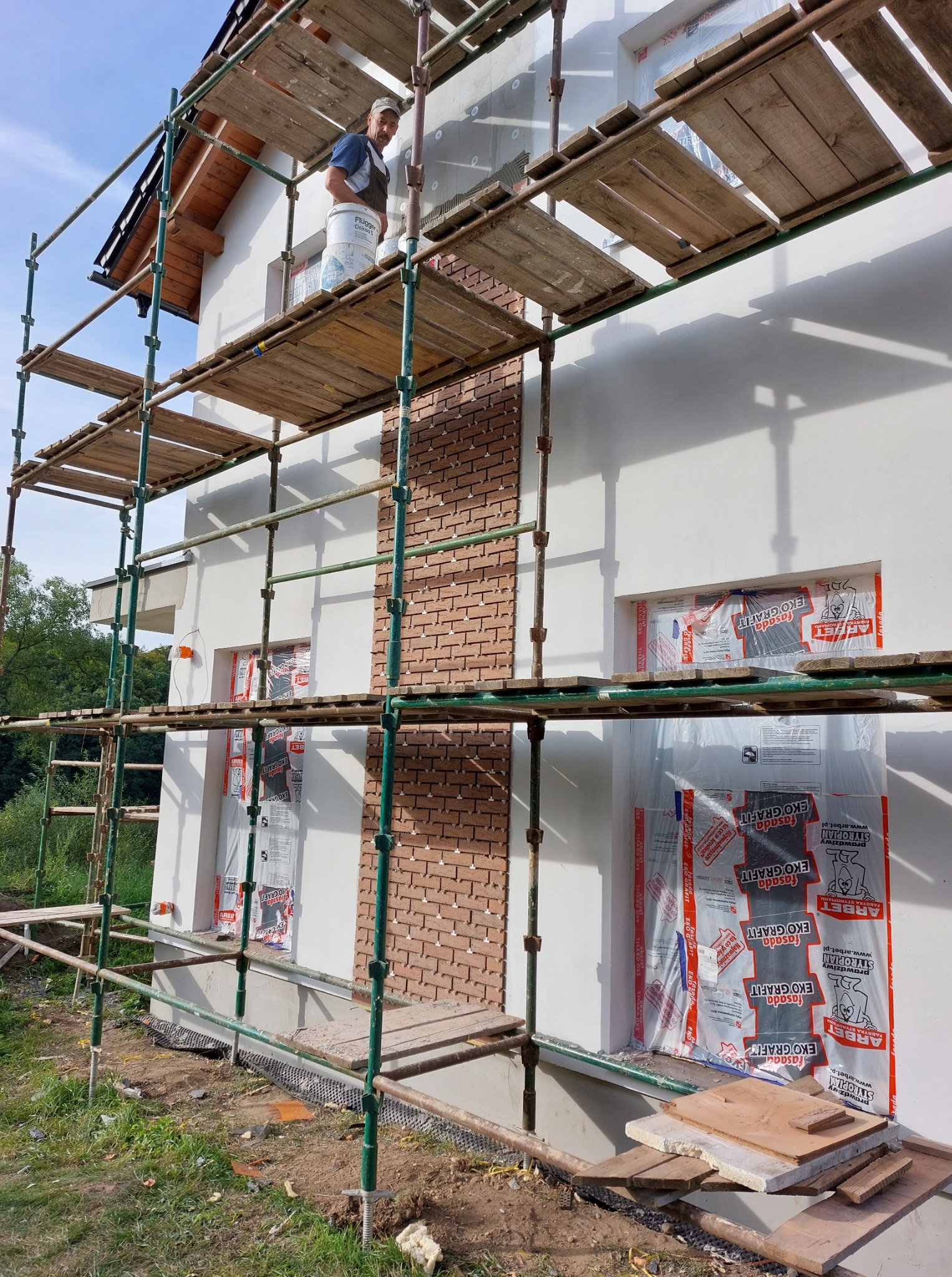 Prace wykończeniowe elewacji domu: murarz na rusztowaniu, obok fragment ściany z cegły klinkierowej, okna zabezpieczone folią. Widoczne narzędzia i materiały budowlane.