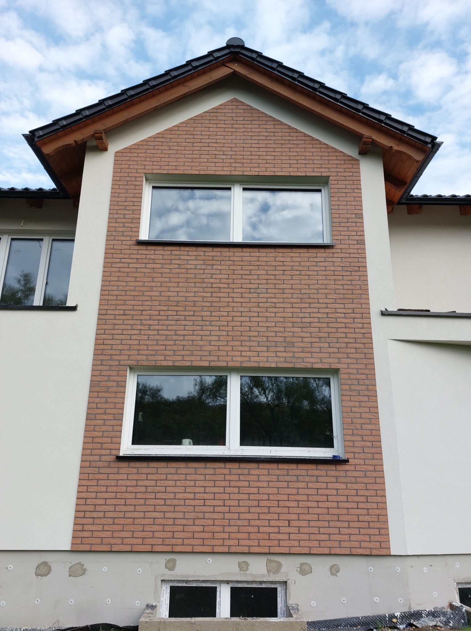 Elewacja domu z dwoma oknami, wykończona cegłą klinkierową w odcieniach brązu i beżu. Widoczne fragmenty ocieplenia fundamentów i okno piwniczne. Dach dwuspadowy z drewnianymi elementami.