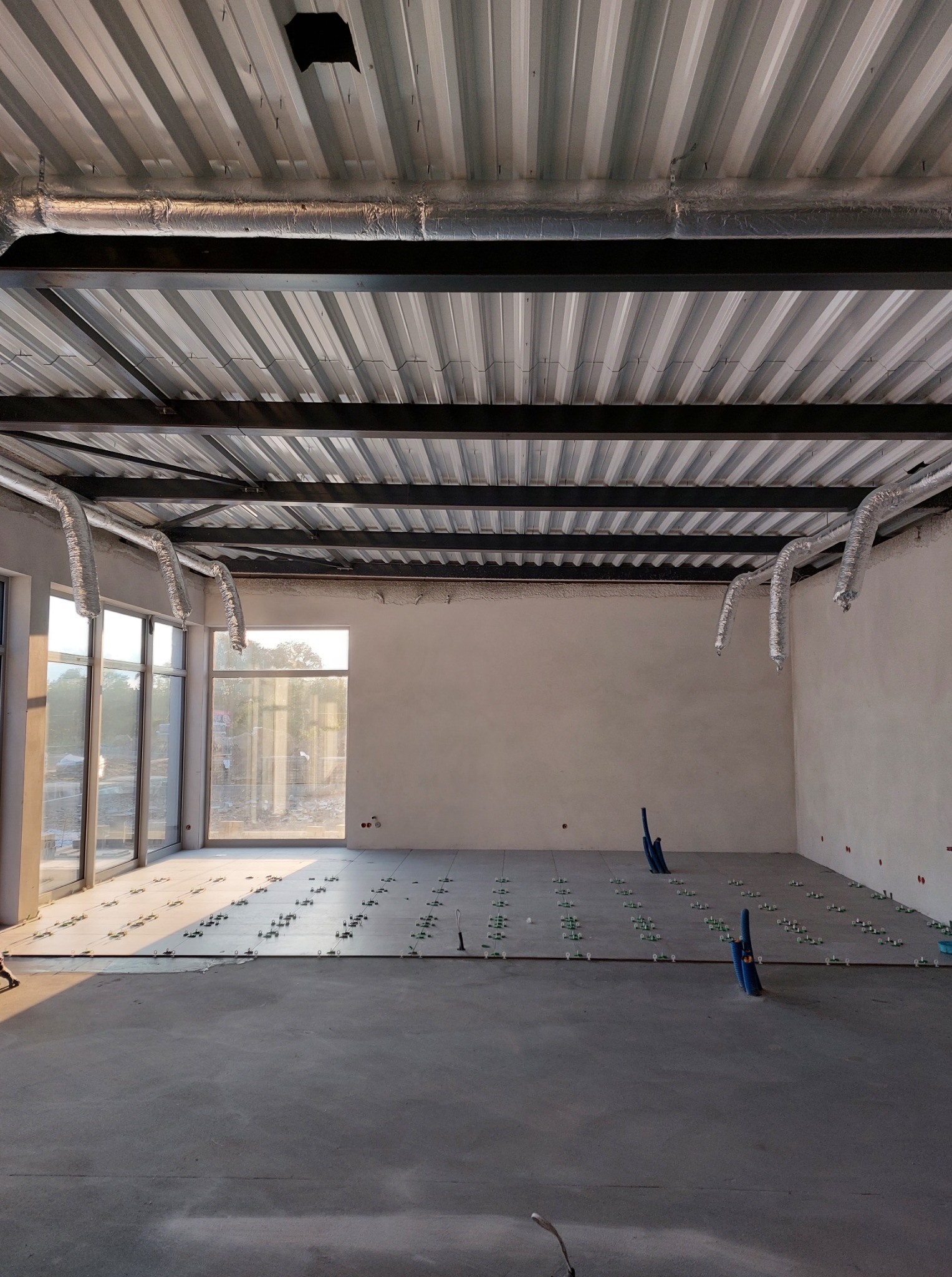 Wnętrze w trakcie prac wykończeniowych: ułożone płytki podłogowe z krzyżykami, widoczne instalacje pod sufitem i okna. Surowy stan, przygotowanie do dalszych prac.