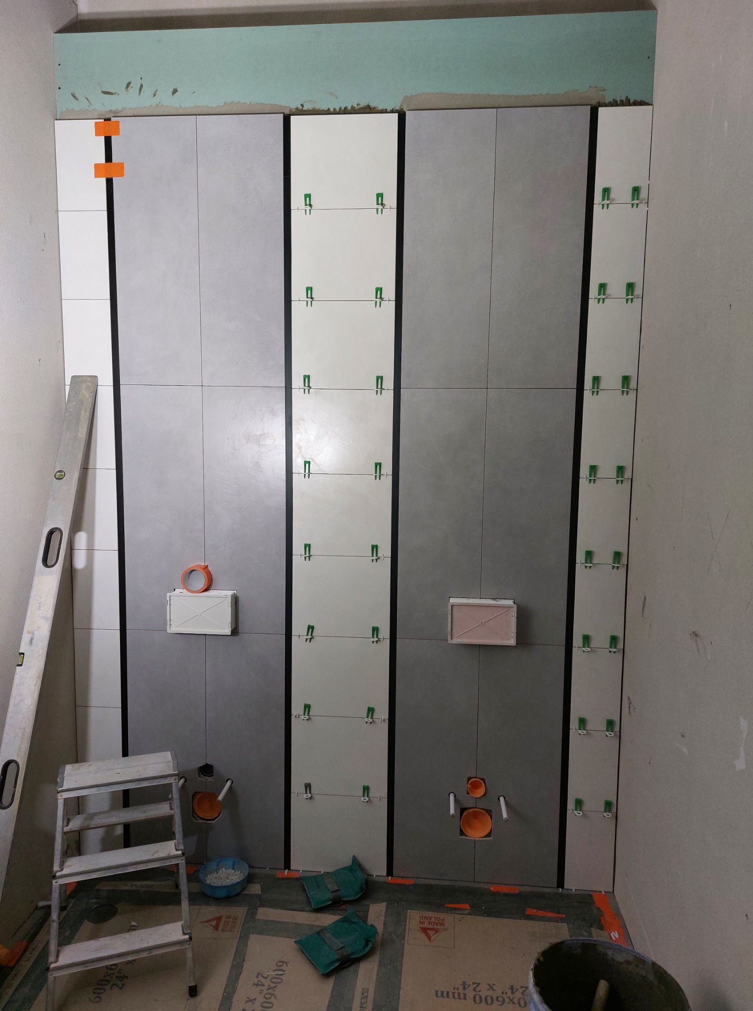 Ściana łazienki w trakcie układania płytek: szare i białe płytki z zielonymi krzyżykami dystansowymi. Widoczne narzędzia glazurnicze i instalacja sanitarna.