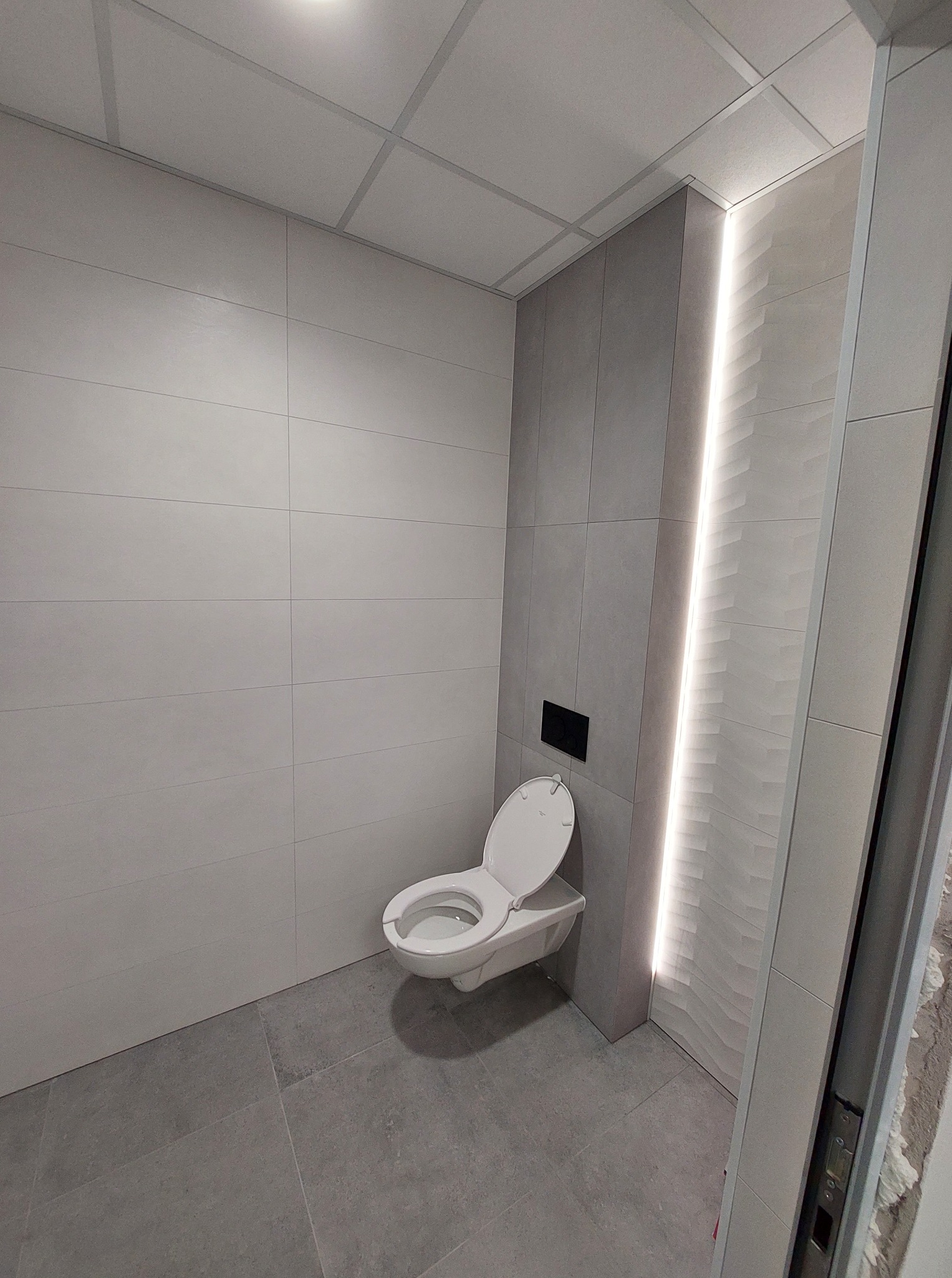 Nowoczesna, wykończona łazienka z podświetleniem LED w pionowym panelu ściennym. Uwagę przyciąga wisząca toaleta i minimalistyczny design płytek w odcieniach szarości.