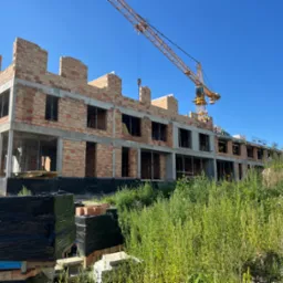 Wznoszenie murów z cegły na budowie wielorodzinnego budynku. Widoczny dźwig budowlany, palety z cegłami oraz bujna roślinność na pierwszym planie.