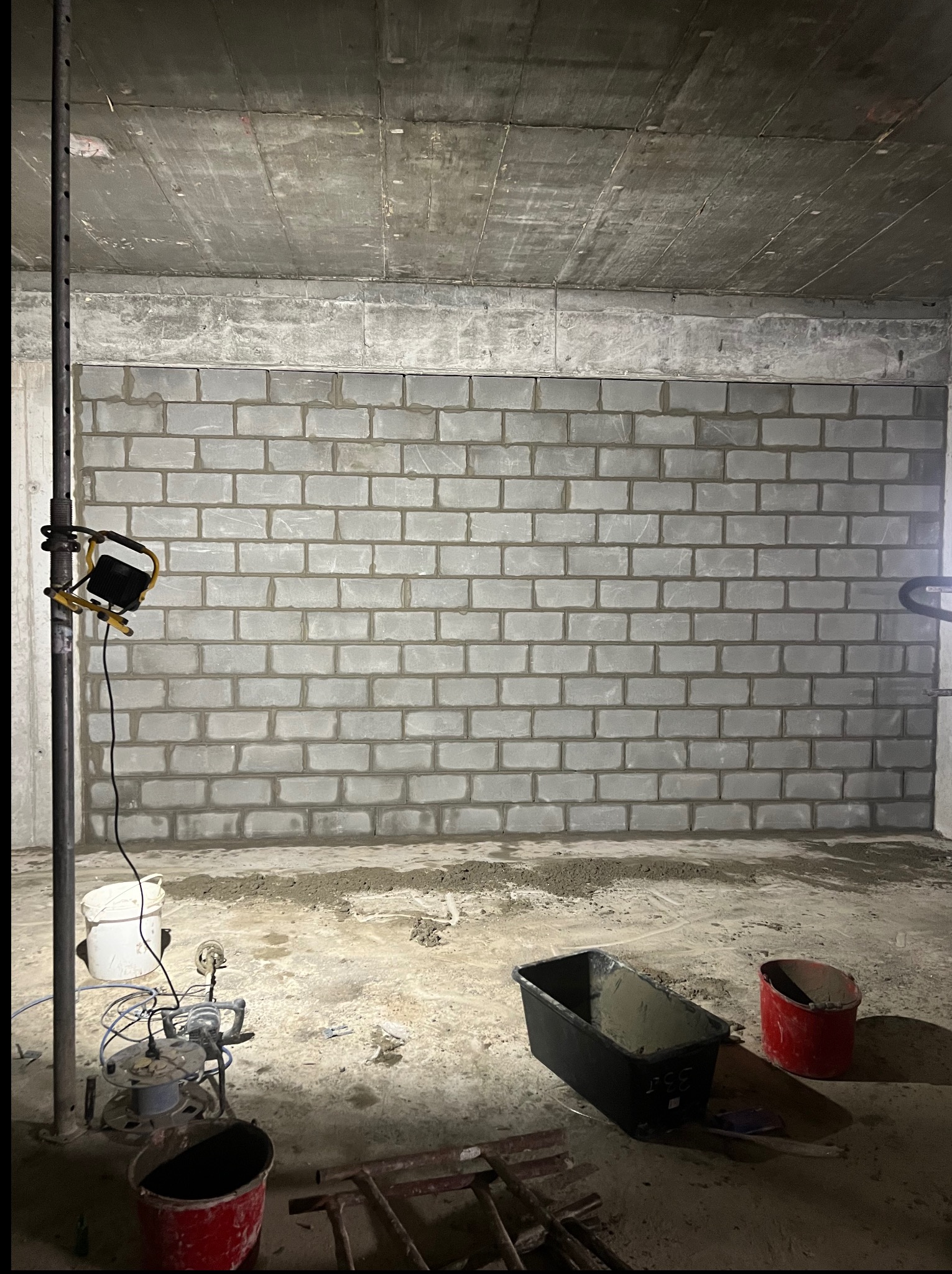 Ściana z szarych bloczków, świeżo wymurowana, widoczne narzędzia i wiadro. Surowy betonowy strop. Oświetlenie budowlane. Etap prac murarskich w piwnicy.
