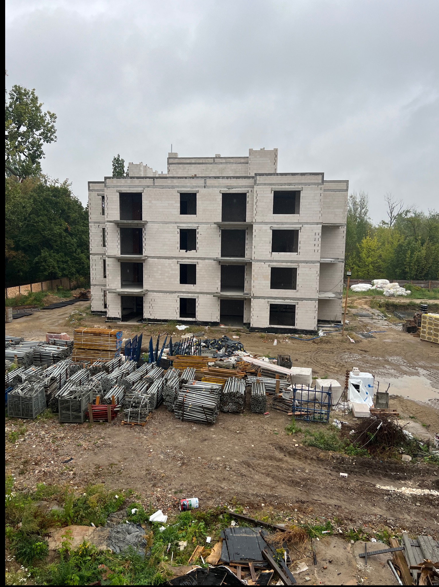 Surowy stan budynku wielorodzinnego z betonu komórkowego, z widocznymi rusztowaniami i materiałami budowlanymi na placu budowy, pochmurny dzień.