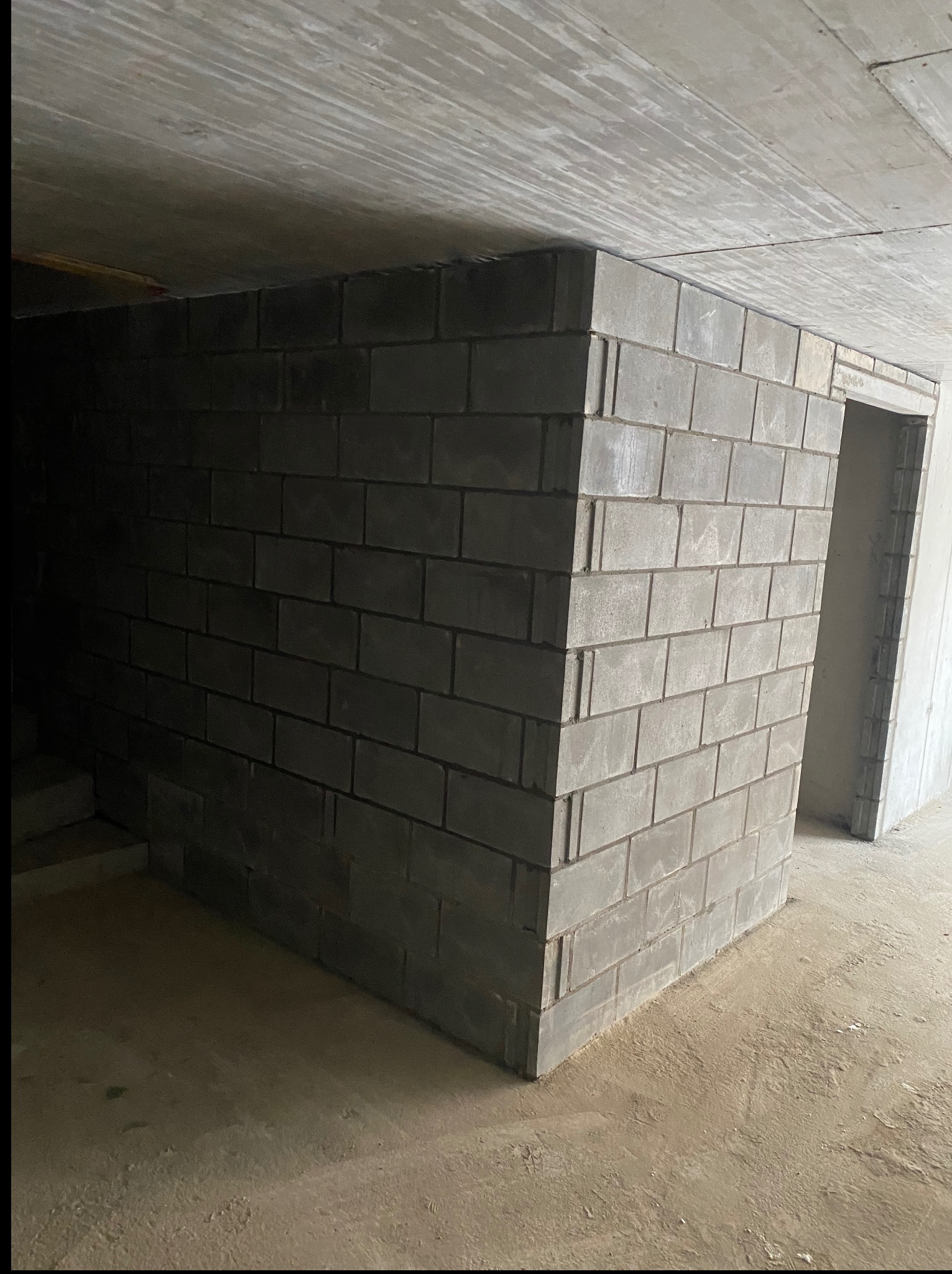 Szara ściana z bloczków betonowych w surowym wnętrzu, widoczny fragment betonowego sufitu i otwór drzwiowy. Prace murarskie w nowym budynku.