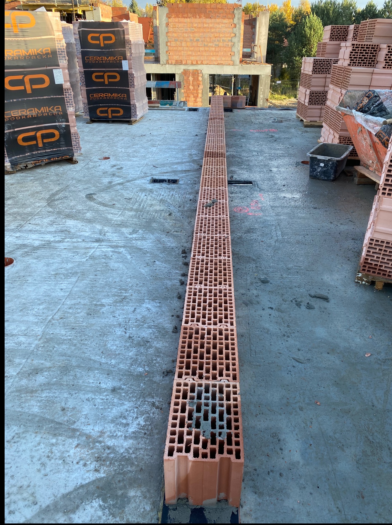 Wstępny etap budowy: Ułożony pierwszy rząd czerwonych, porowatych pustaków ceramicznych na fundamencie, w tle palety z materiałem budowlanym i fragment konstrukcji domu.