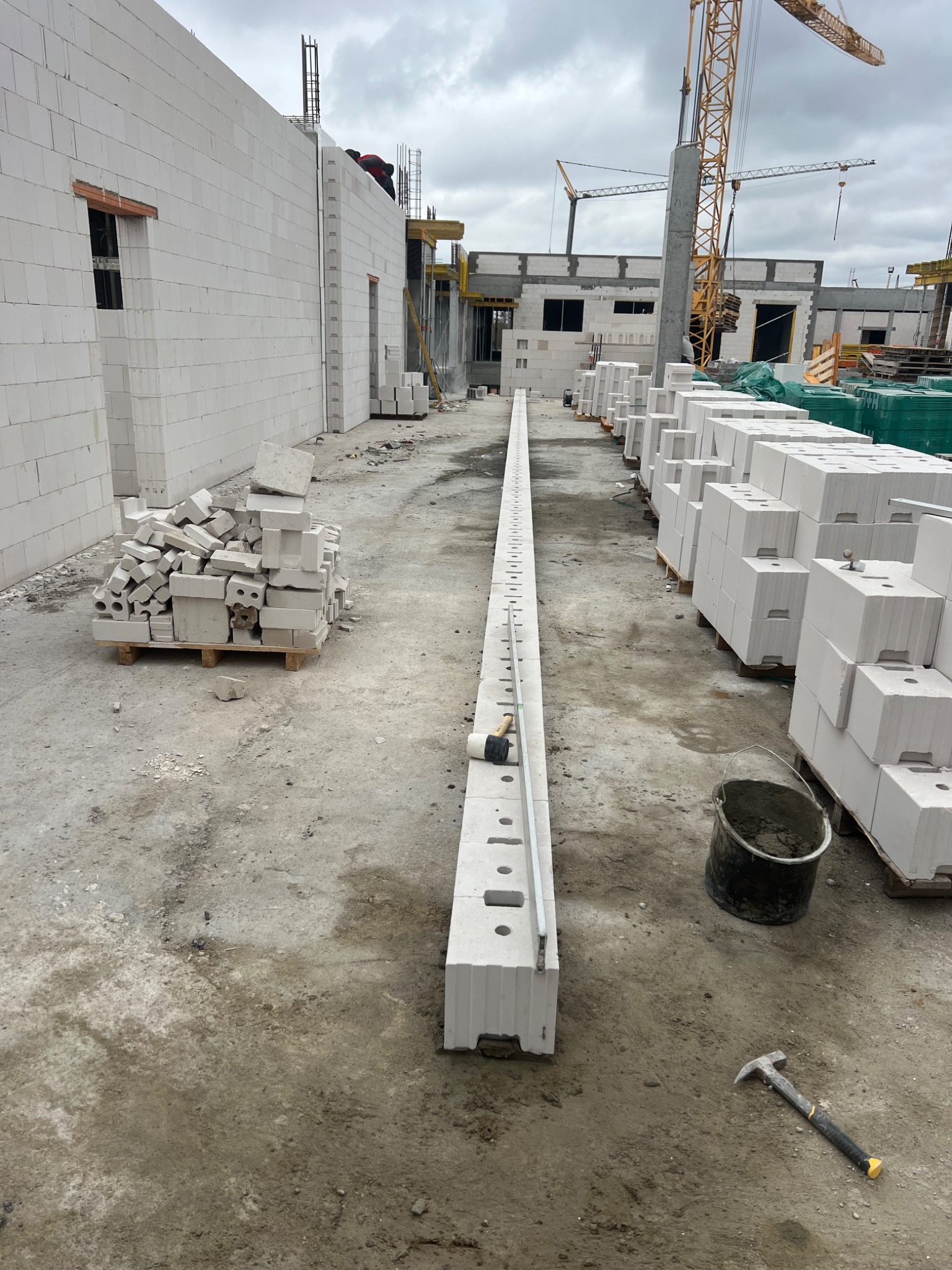 Budowa domu z białych bloczków: fragment ściany z oknem, ułożone bloczki, narzędzia murarskie, żuraw w tle. Widok na plac budowy w pochmurny dzień.