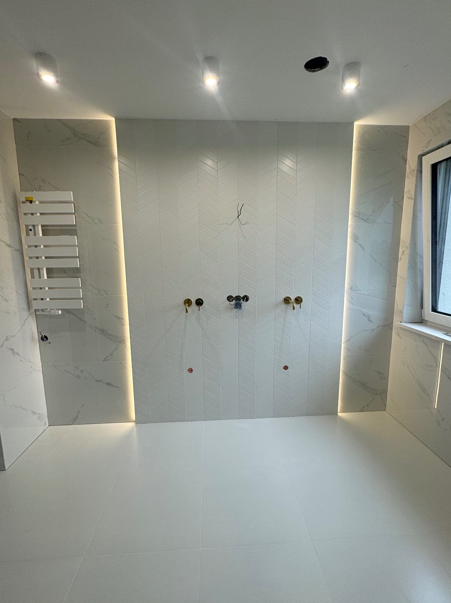 Biała łazienka z marmurowymi płytkami, oświetleniem LED i złotą armaturą. Ściana z wzorem w jodełkę. Grzejnik drabinkowy po lewej. Minimalistyczny design.