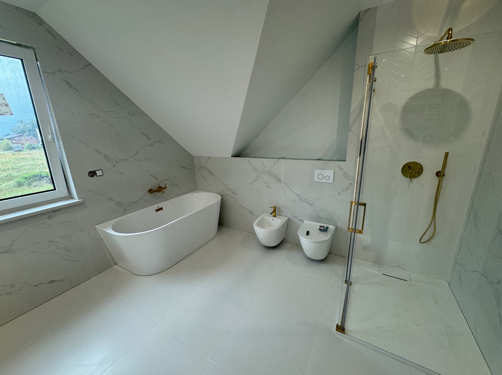 Nowoczesna łazienka na poddaszu z wanną, bidetem, toaletą i kabiną prysznicową ze złotą armaturą. Marmurowe płytki na ścianach i jasna podłoga.