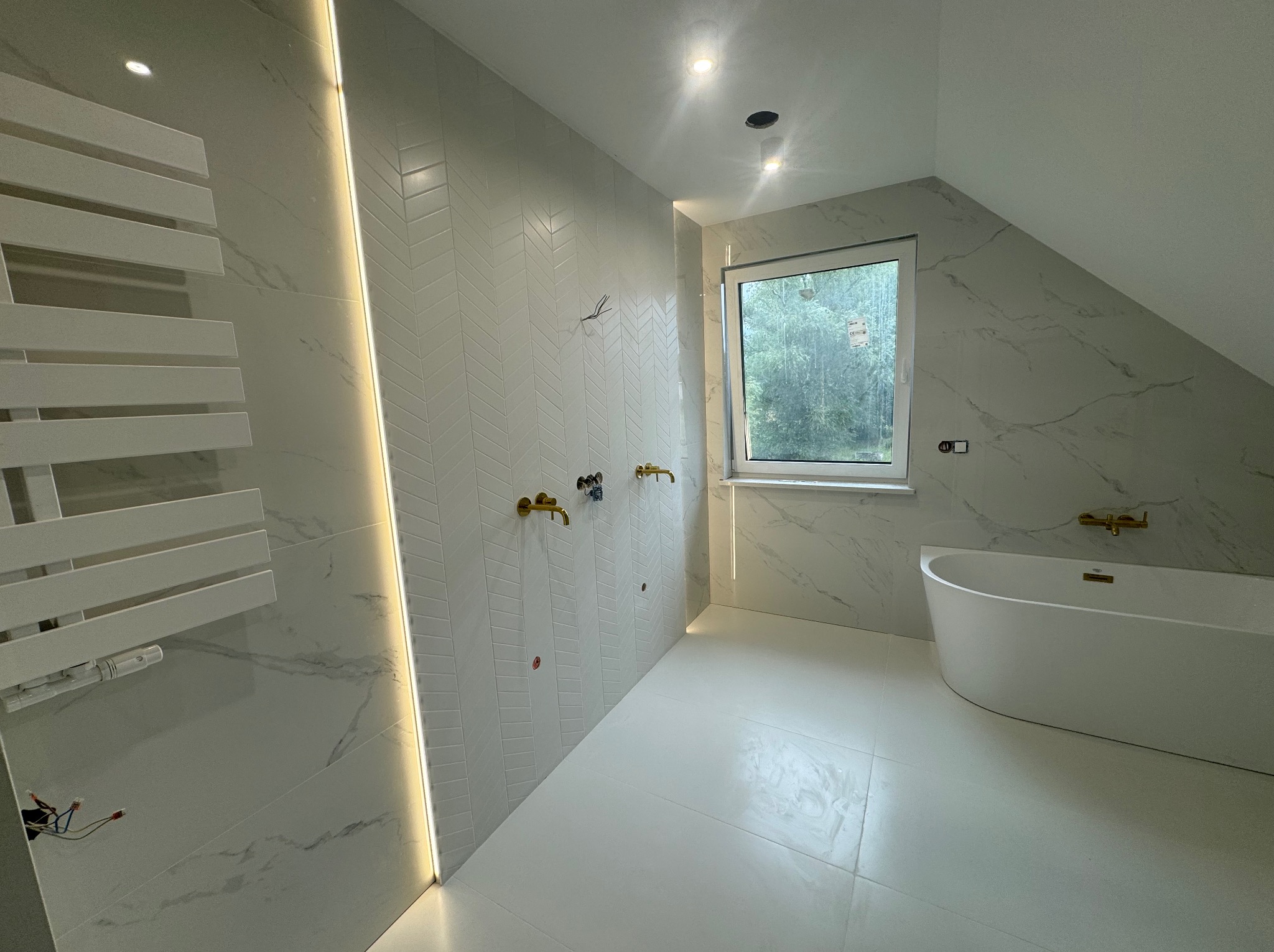 Elegancka łazienka na poddaszu z białą wanną, marmurowymi płytkami i złotą armaturą. Uwagę zwraca oświetlenie LED i wzór jodełki na ścianie.