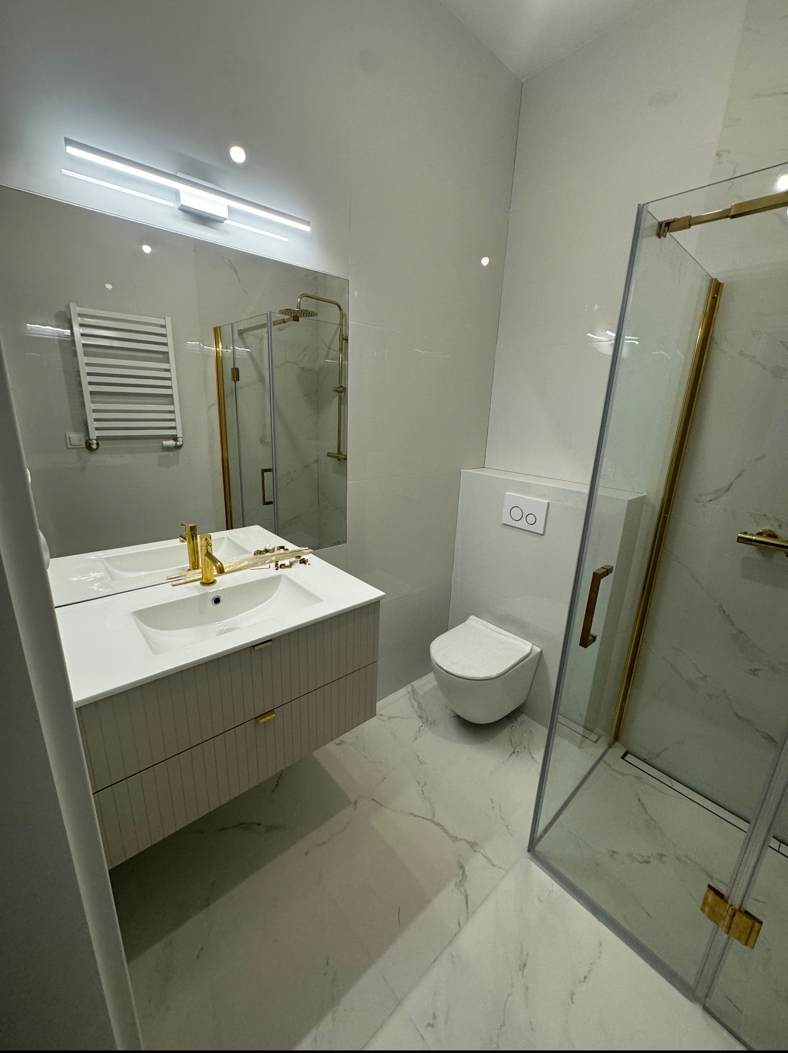 Nowoczesna łazienka w bieli z akcentami złota: kabina prysznicowa, umywalka z szafką, toaleta podwieszana, lustro z oświetleniem. Marmurowe płytki podłogowe.