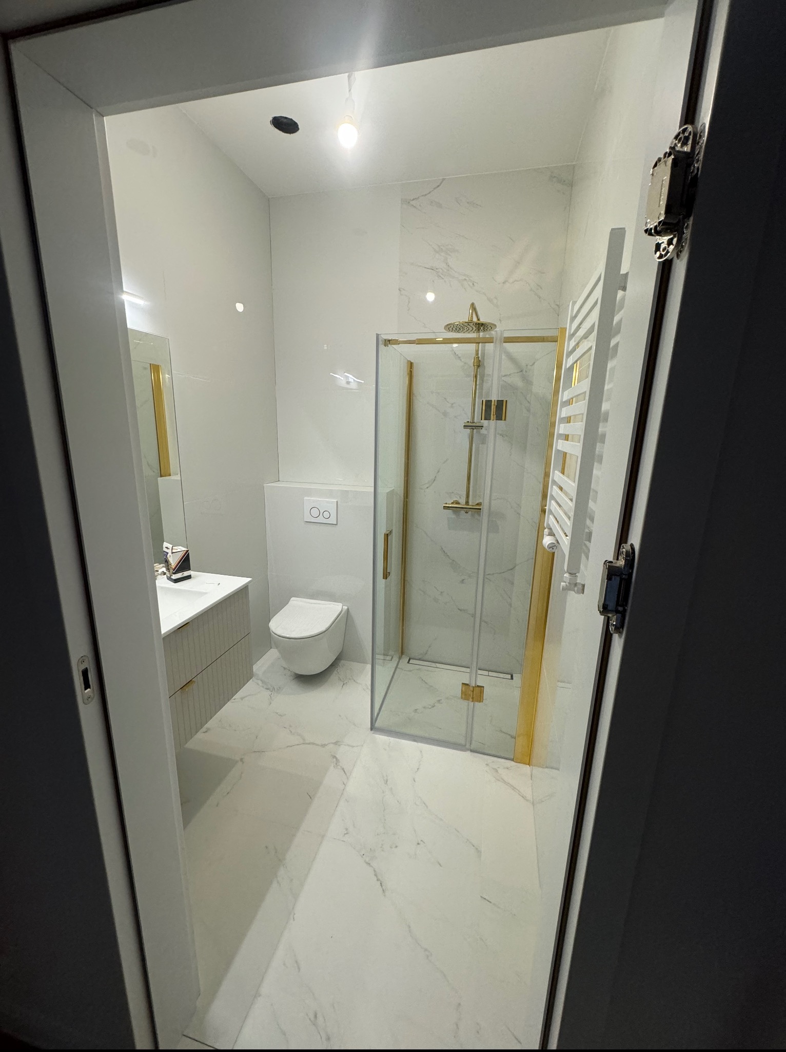 Nowoczesna łazienka z białymi marmurowymi płytkami, złotymi akcentami w kabinie prysznicowej i armaturze, widok zza uchylonych drzwi.