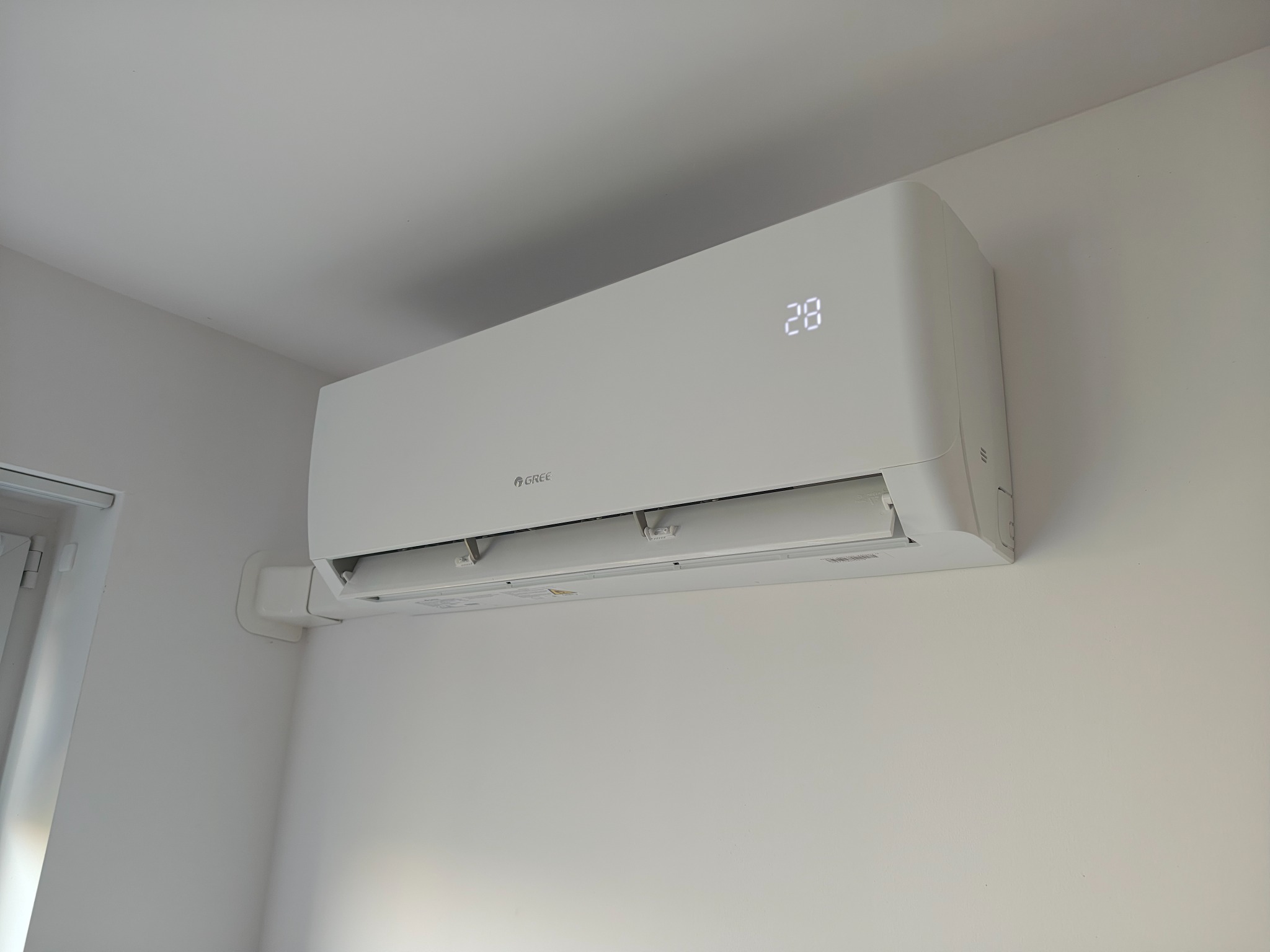 Biała klimatyzacja Gree zamontowana na ścianie w jasnym pomieszczeniu, wyświetlająca temperaturę 28 stopni Celsjusza. Widoczny fragment okna po lewej stronie.