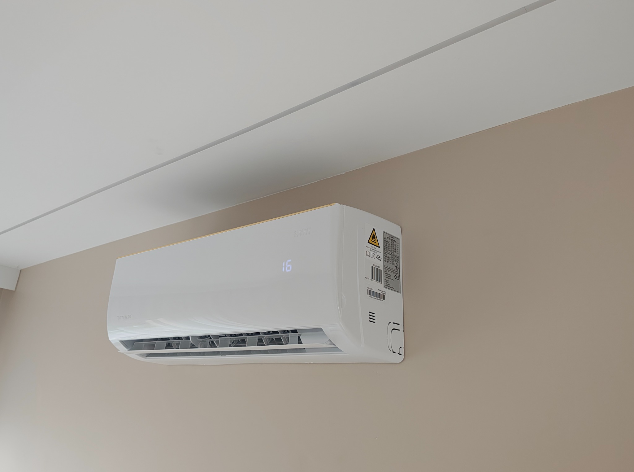 Nowoczesny biały klimatyzator na beżowej ścianie, z wyświetlaczem temperatury 16 stopni, widoczny fragment sufitu. Minimalistyczny design.