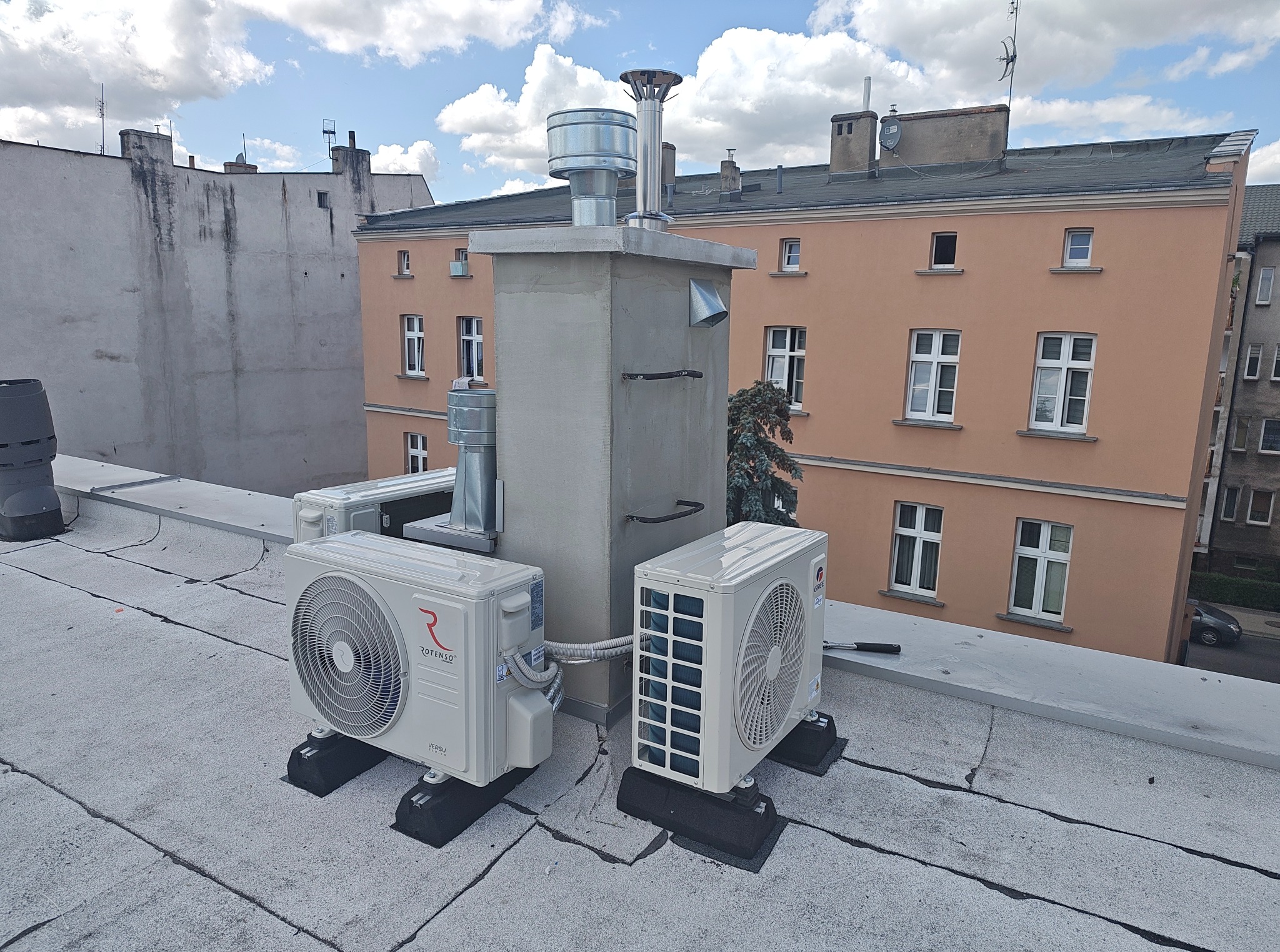 Dwa klimatyzatory marki Rotenso i Gree na dachu budynku, obok komina wentylacyjnego. Widoczne elementy instalacji i fragmenty sąsiednich budynków.