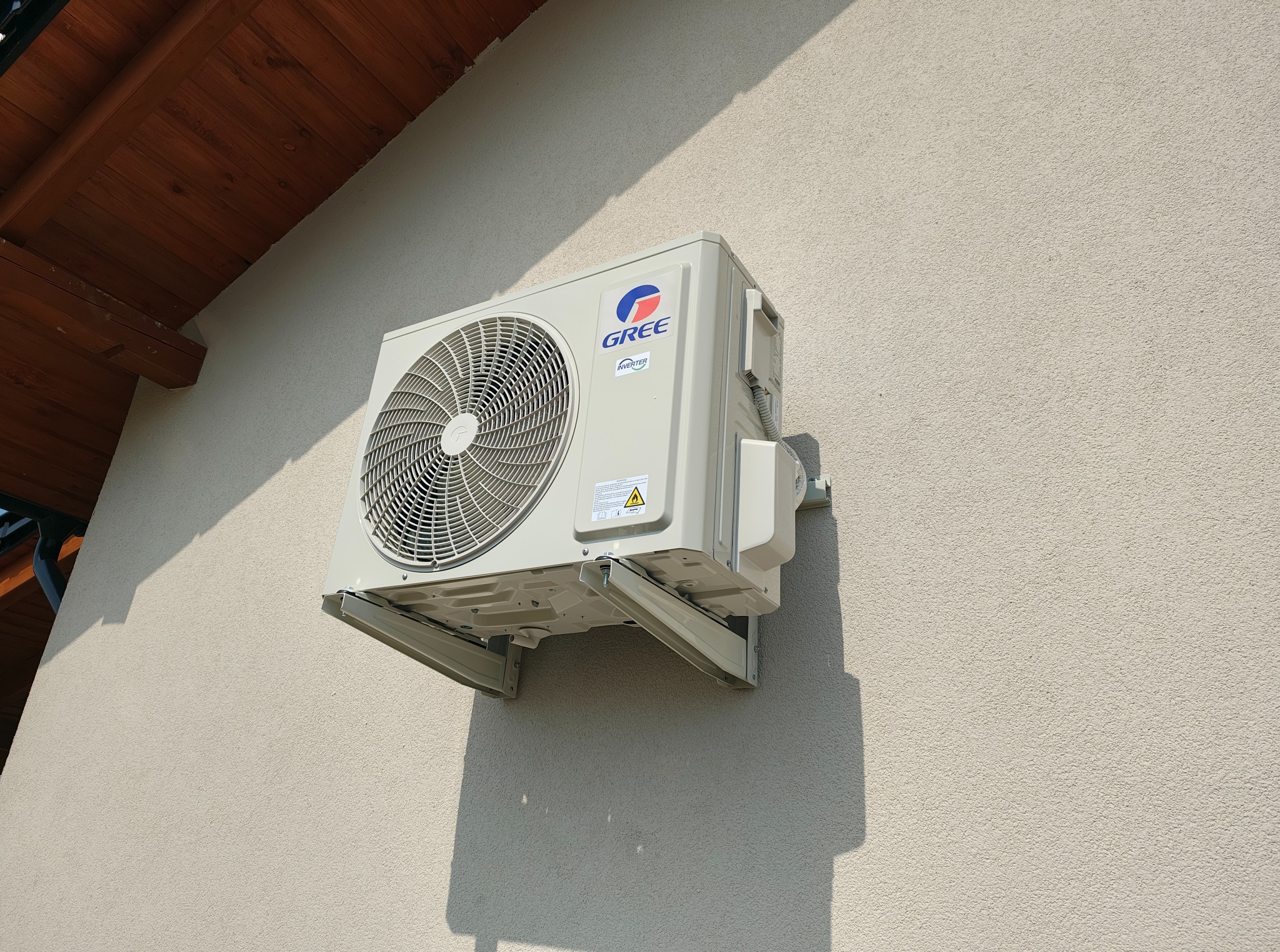Zamontowana jednostka zewnętrzna klimatyzacji Gree na elewacji domu, widoczna z boku, z fragmentem dachu w tle. Jasne światło słoneczne.