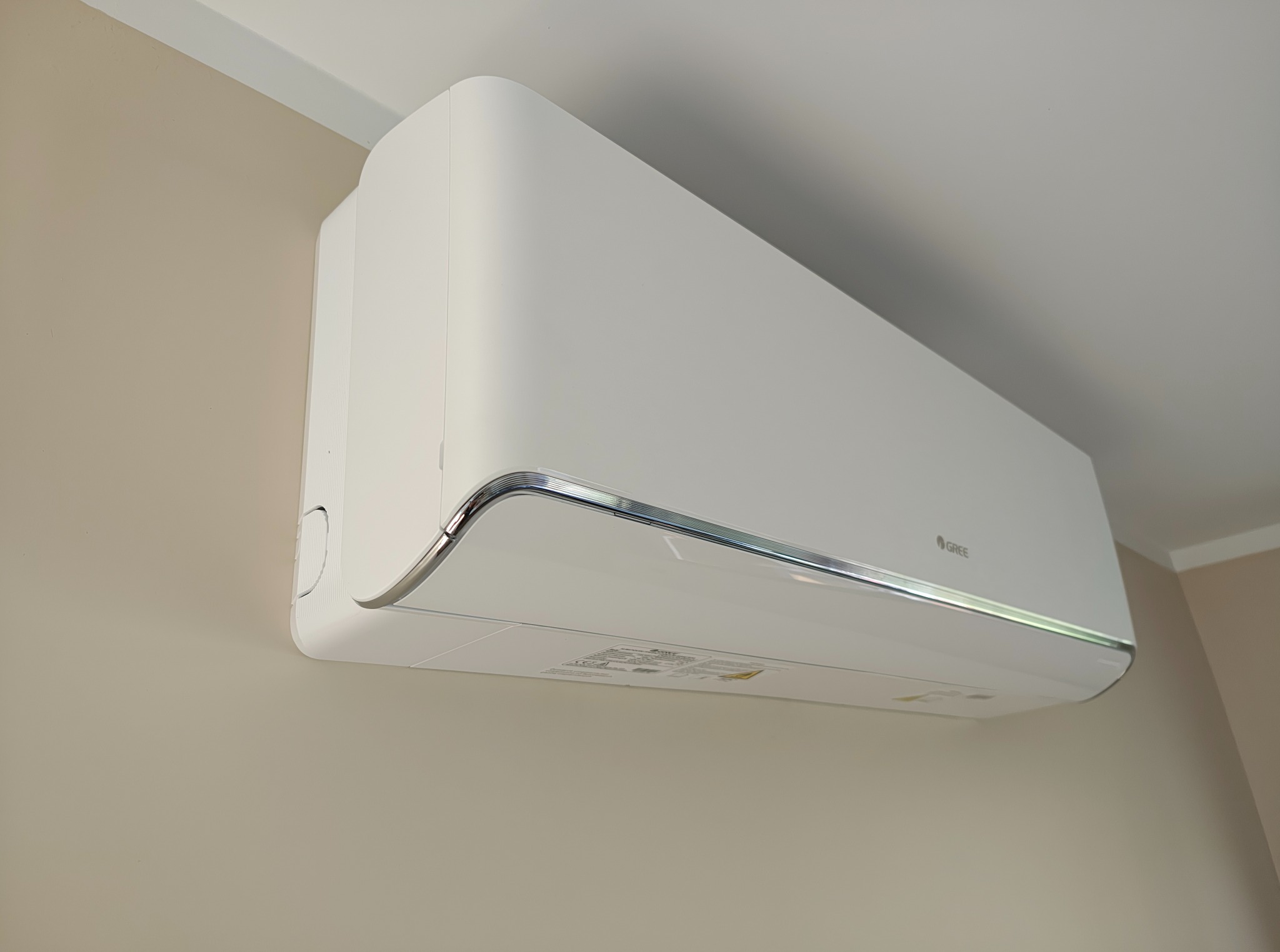 Biała klimatyzacja GREE, zamontowana na beżowej ścianie w jasnym wnętrzu. Widoczny fragment sufitu i listwy przysufitowej. Ujęcie z dołu.