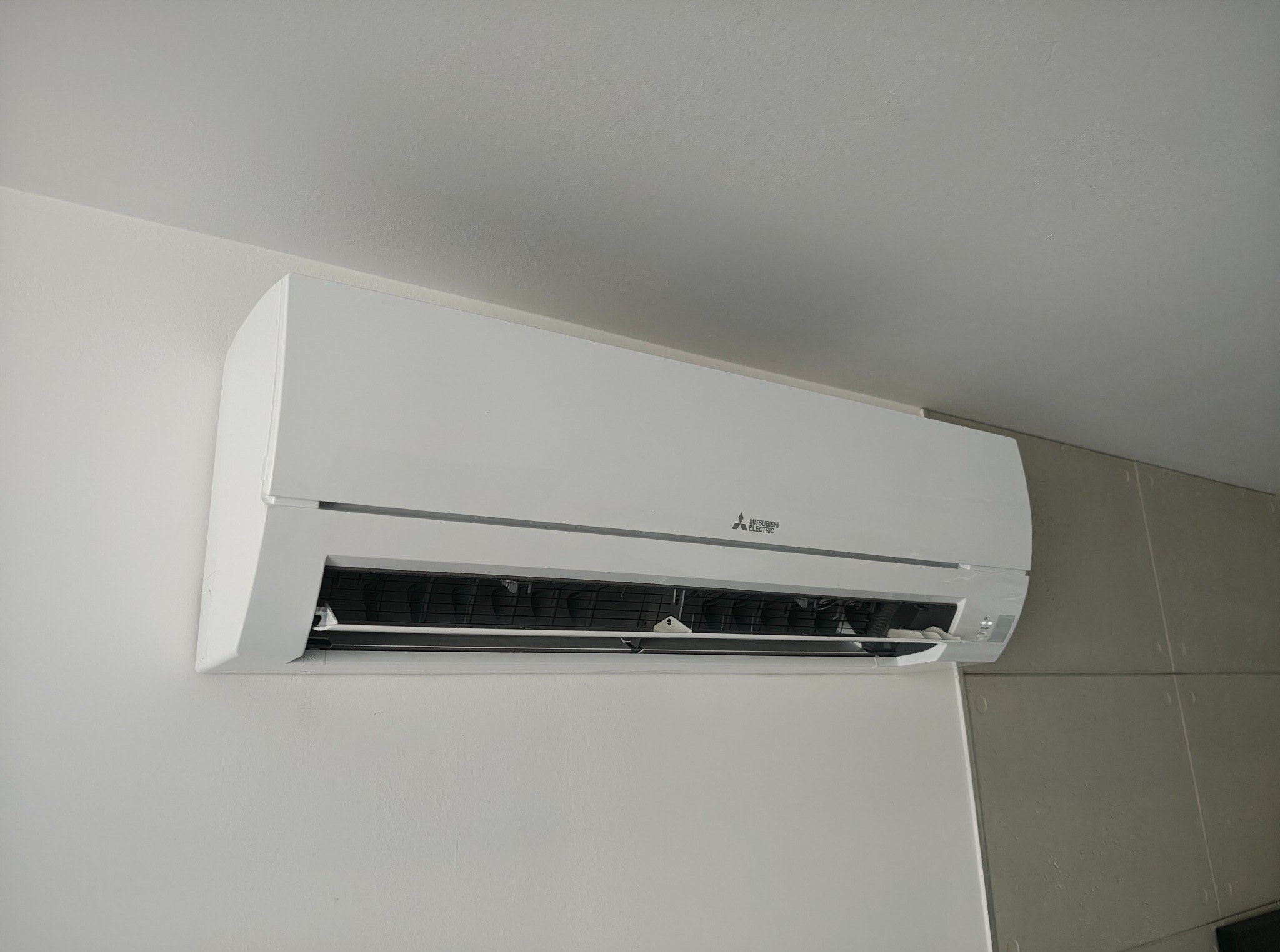 Biała klimatyzacja Mitsubishi Electric zamontowana na jasnej ścianie. Widoczny fragment sufitu i sąsiedniej ściany wykończonej płytkami.