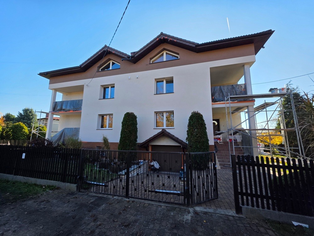 Dwukondygnacyjny dom z beżową elewacją i brązowym wykończeniem dachu, widoczny zza czarnego ogrodzenia. Rusztowania na balkonie sugerują prace wykończeniowe.