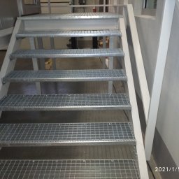 Tatorex - Metalowe schody kratowe z białymi poręczami w hali przemysłowej, widok z dołu na stopnie.