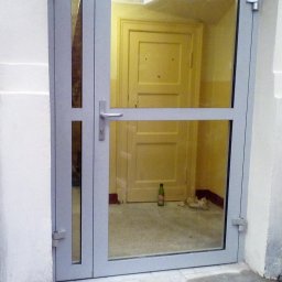 Tatorex - Szare aluminiowe drzwi wejściowe z szybą, widoczne wnętrze z żółtymi drzwiami i butelką na podłodze.