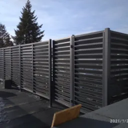 Nowoczesna, pozioma, metalowa przesłona tarasowa w kolorze grafitowym, z widocznymi szczelinami, zapewniająca prywatność na tle zimowego krajobrazu z drzewami iglastymi i resztkami śniegu.