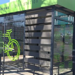 Tatorex - Zadaszona stacja rowerowa z elementami z kompozytu, z grafiką roweru na szybie i napisem 'stacja rowerowa | bike station' w zielonym kolorze.