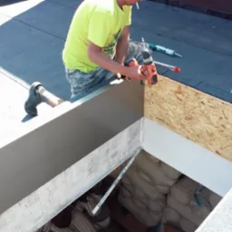 Pracownik w żółtej koszulce w trakcie montażu obudowy włazu dachowego z wykorzystaniem wkrętarki na dachu pokrytym papą. W tle widoczne worki z materiałem budowlanym.