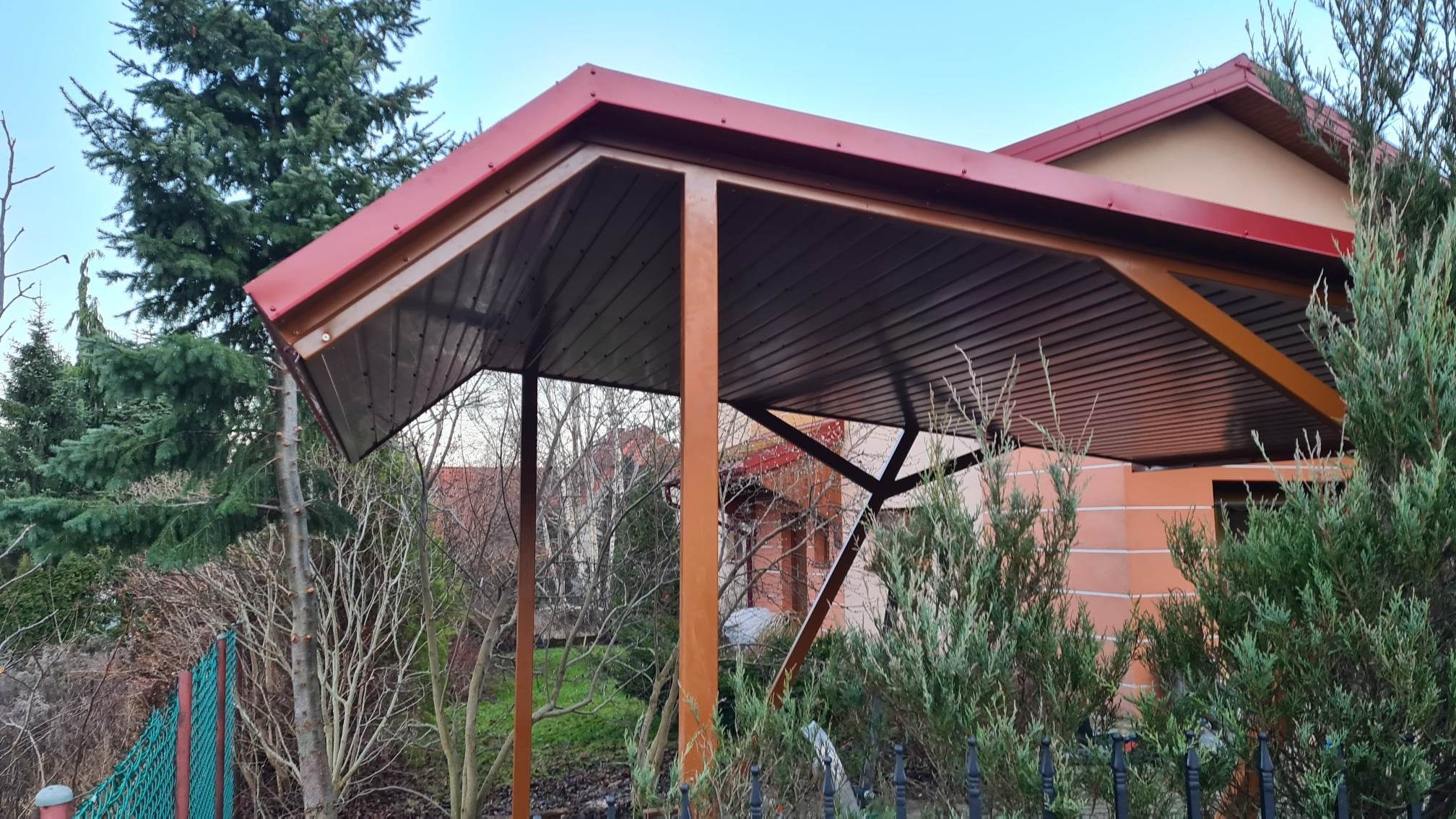 Metalowa konstrukcja z czerwonym dachem, prawdopodobnie zadaszenie tarasu lub wiaty, widoczna z perspektywy ogrodu, z elementami roślinności i ogrodzenia w tle.