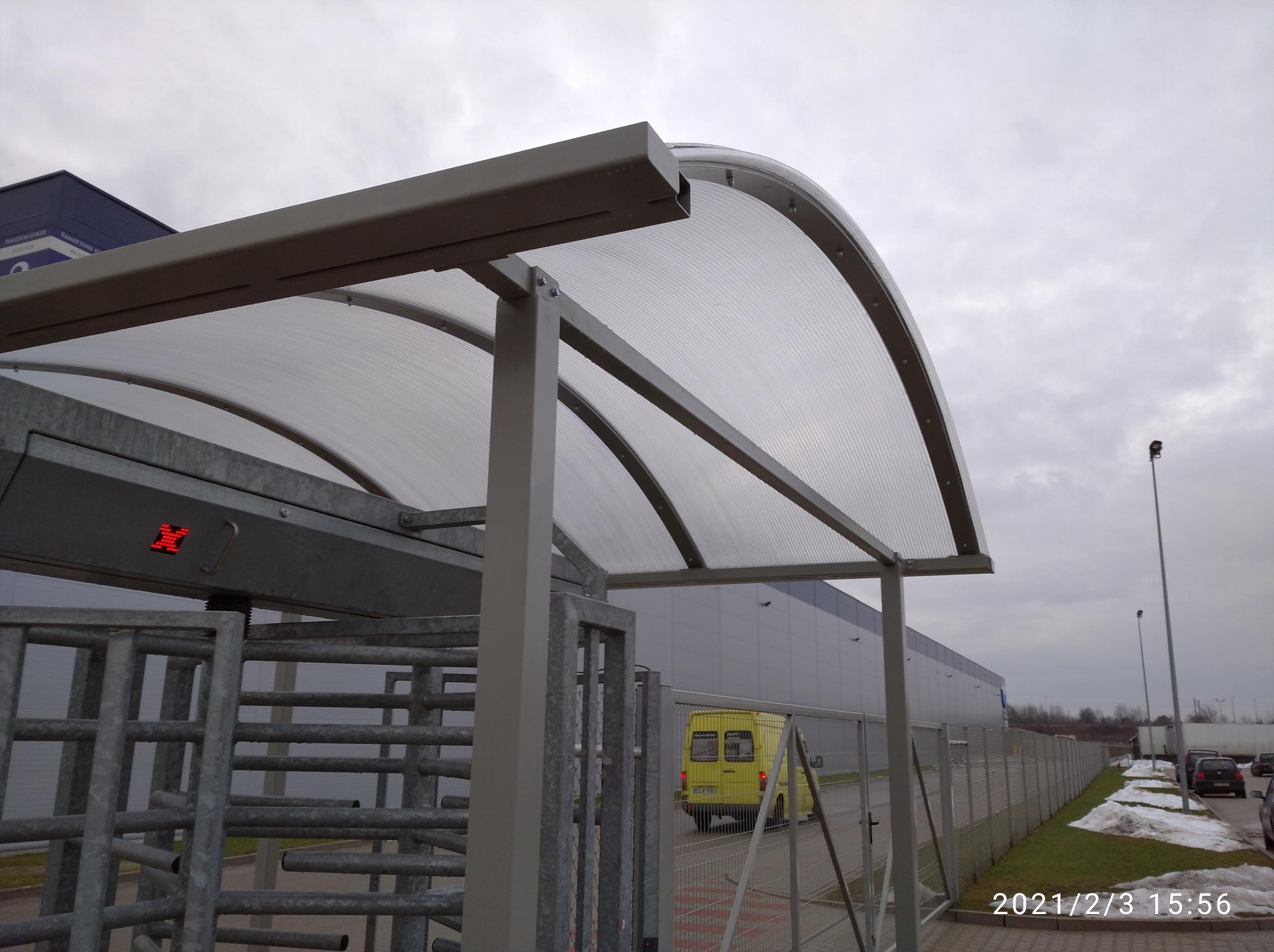 Zewnętrzne przejście z kołowrotami i zadaszeniem z poliwęglanu przy budynku przemysłowym, widoczny żółty samochód dostawczy w tle.