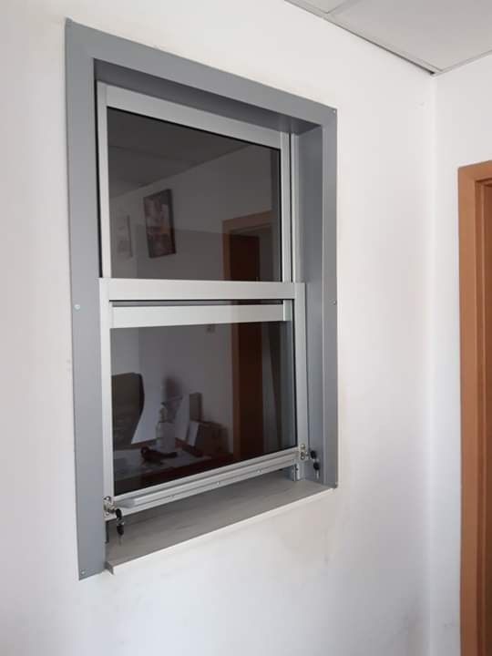 Okno pionowe przesuwne z ramą aluminiową w kolorze szarym, widoczne zamki zabezpieczające na dolnej krawędzi, odbicie wnętrza pomieszczenia w szybie.