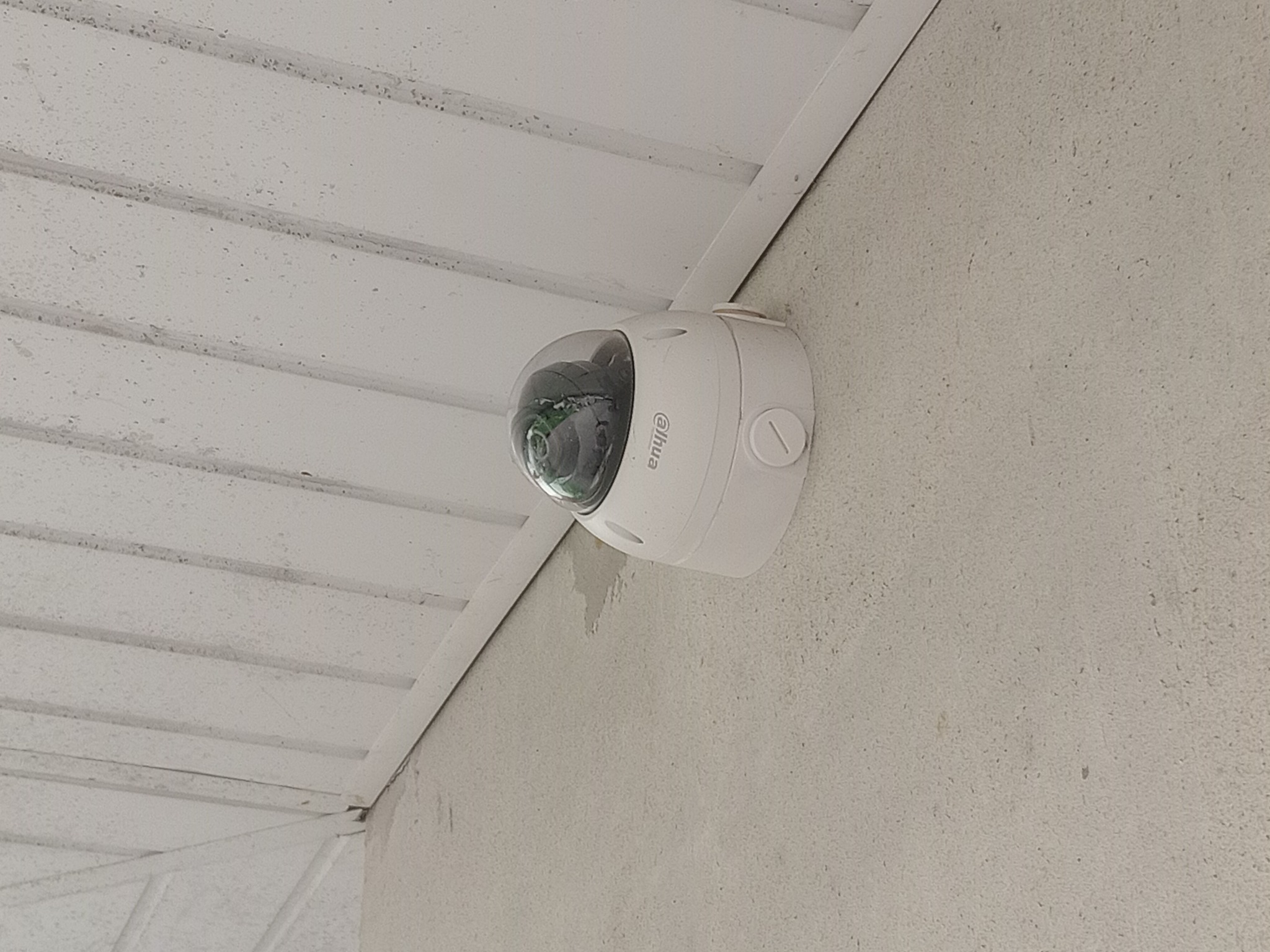 Biała kamera kopułowa Dahua zamontowana pod kątem na jasnej ścianie wewnątrz budynku, widoczne elementy konstrukcyjne sufitu.