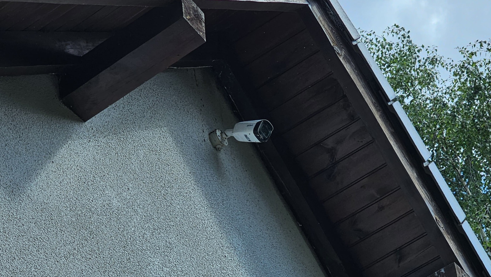 Zewnętrzna kamera monitoringu, biała, zamontowana pod okapem budynku z widoczną fakturą elewacji i fragmentem drzewa. Kątowe ujęcie.