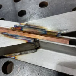 Precyzyjne spawy łączące metalowe profile pod kątem, widoczne tęczowe przebarwienia termiczne, całość na perforowanym stole spawalniczym.