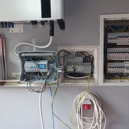 JM Energia Jakub Plechowski - Rozdzielnice elektryczne z otwartymi drzwiczkami, widoczne kable, bezpieczniki i inne komponenty. Na ścianie zamontowany jest inwerter fotowoltaiczny.