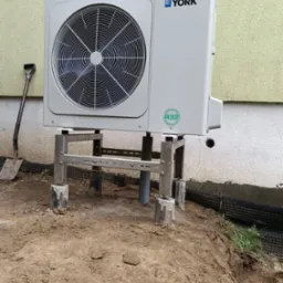 Zewnętrzna jednostka pompy ciepła York, zamontowana na metalowej konstrukcji. Widoczna ściana budynku z otworem, obok leży łopata. Montaż w trakcie prac ziemnych.
