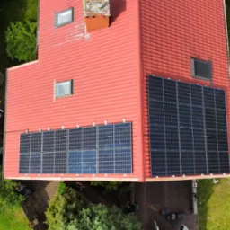 Widok z góry na dom z czerwoną dachówką i panelami fotowoltaicznymi. Panele zamontowane na dwóch połaciach dachu, otoczony zielenią i częściowo brukowanym terenem.