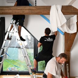 JM Energia Jakub Plechowski - Montaż klimatyzacji w domu z antresolą: trzech monterów na różnych poziomach, jeden na drabinie, drugi na belce, trzeci na podłodze, widoczne narzędzia i elementy instalacji.