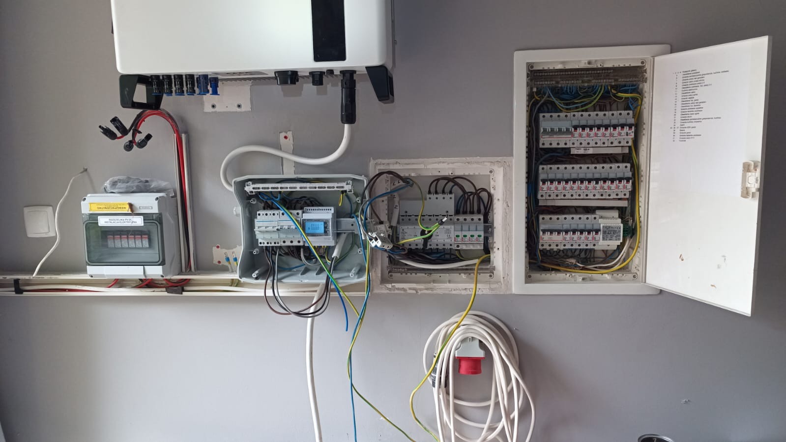Rozdzielnice elektryczne z otwartymi drzwiczkami, widoczne kable, bezpieczniki i inne komponenty. Na ścianie zamontowany jest inwerter fotowoltaiczny.