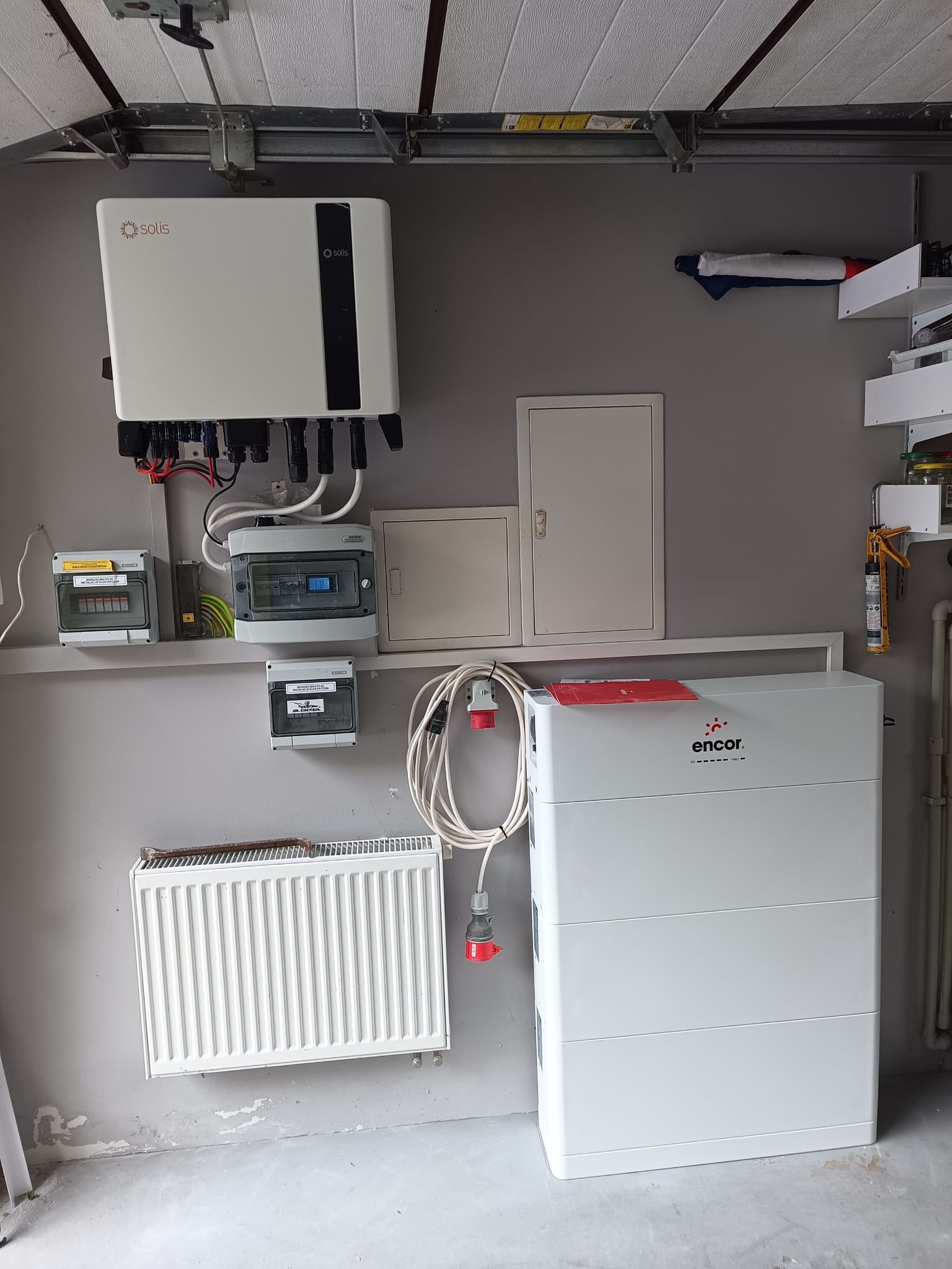 Solis falownik i magazyn energii Encor w garażu, widoczne zabezpieczenia, instalacja elektryczna, grzejnik, szare ściany.