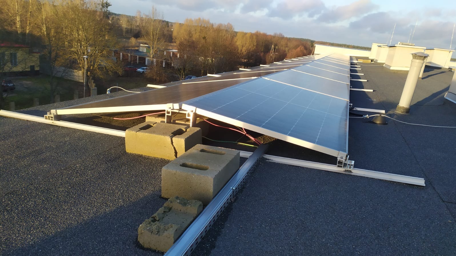 Panele fotowoltaiczne na płaskim dachu budynku, wsparte na betonowych blokach, z widocznymi przewodami. Słoneczny dzień, w tle drzewa i budynki.