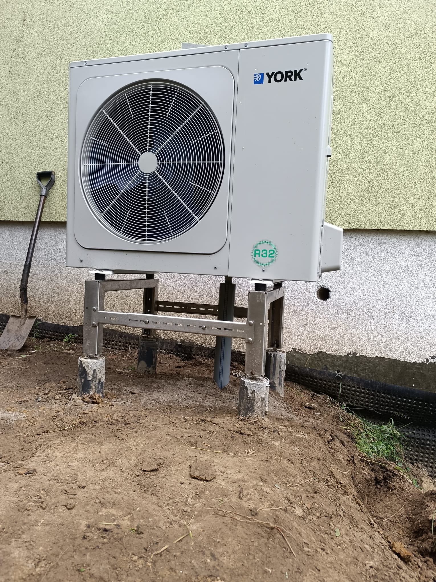 Zewnętrzna jednostka pompy ciepła York, zamontowana na metalowej konstrukcji. Widoczna ściana budynku z otworem, obok leży łopata. Montaż w trakcie prac ziemnych.