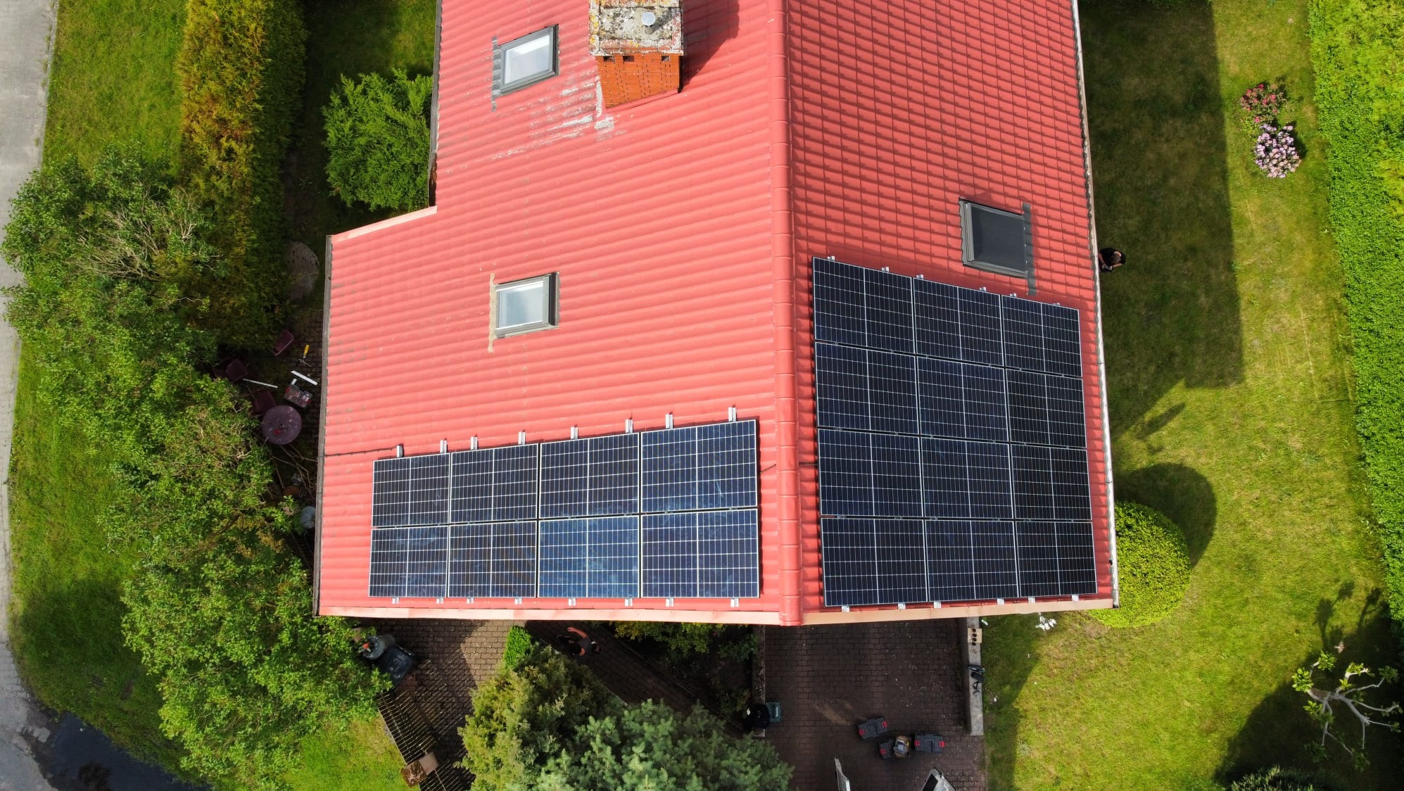 Widok z góry na dom z czerwoną dachówką i panelami fotowoltaicznymi. Panele zamontowane na dwóch połaciach dachu, otoczony zielenią i częściowo brukowanym terenem.
