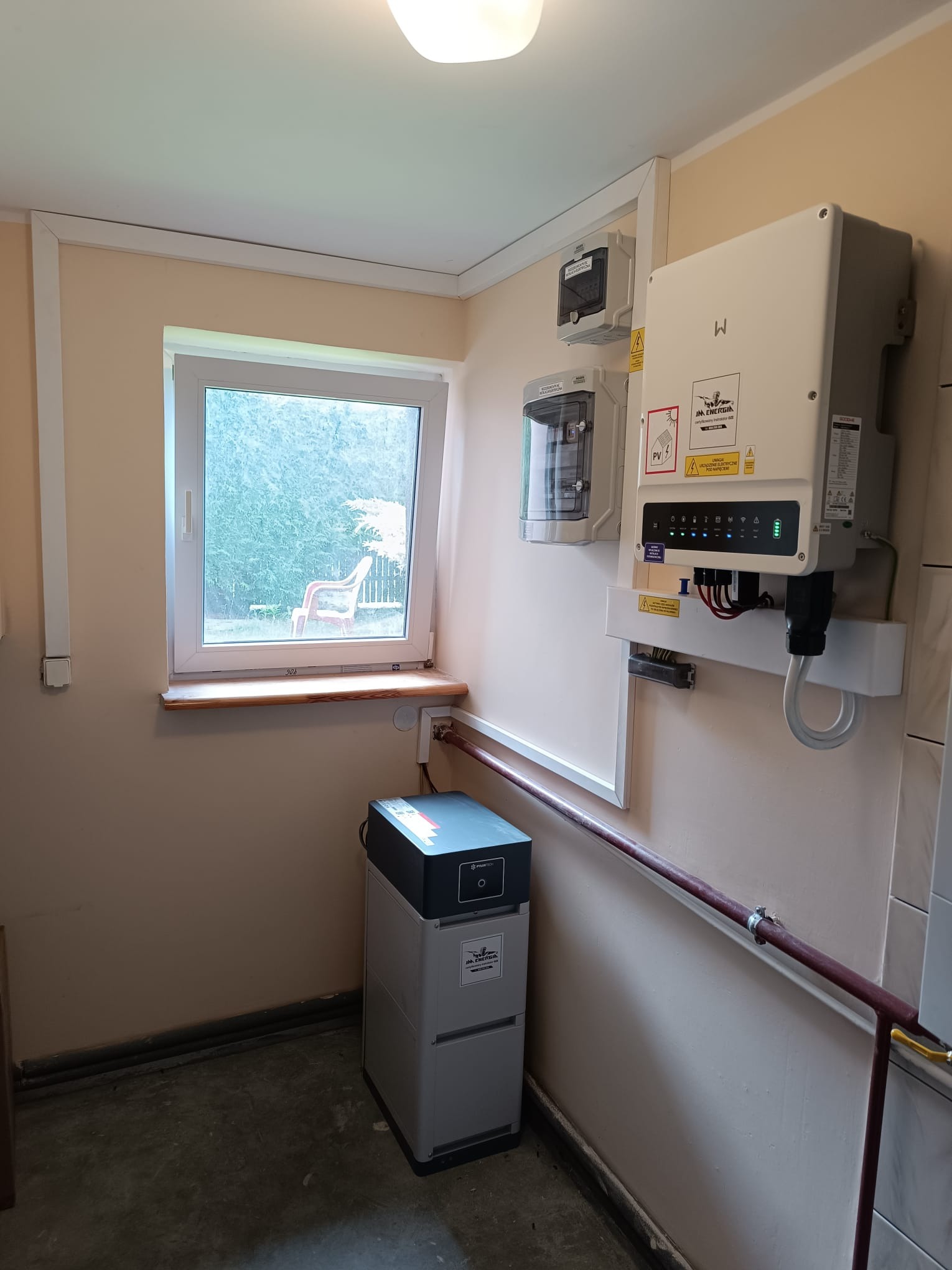 Wnętrze pomieszczenia z zamontowanym inwerterem fotowoltaicznym na ścianie, obok okna i z akumulatorem energii na podłodze, instalacja elektryczna widoczna.