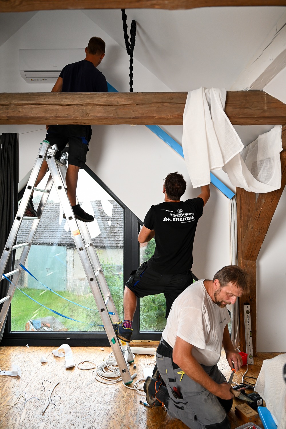 Montaż klimatyzacji w domu z antresolą: trzech monterów na różnych poziomach, jeden na drabinie, drugi na belce, trzeci na podłodze, widoczne narzędzia i elementy instalacji.